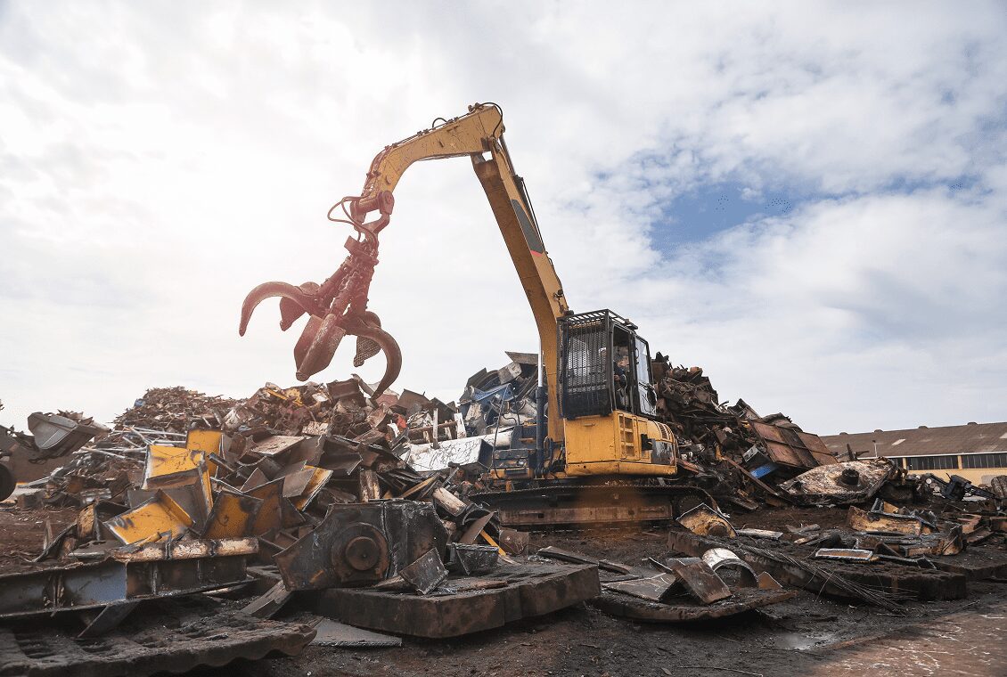 A yellow excavator sorting scrap metal outdoors under a cloudy sky.
